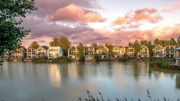 A row of modern, multi-story buildings with large windows, situated by a calm lake, reflecting the sky. - Modern Cotswolds lakeside retreat, spa and pool (Somerford Keynes)