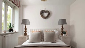 A bedroom with a white bed, two bedside tables with lamps, a heart-shaped wall decoration, and a window with a view of greenery. - Modern Cotswolds lakeside retreat, spa and pool (Somerford Keynes)
