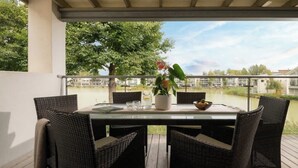 A covered outdoor dining area with wicker chairs and a table, overlooking a body of water and buildings. - Modern Cotswolds lakeside retreat, spa and pool (Somerford Keynes)