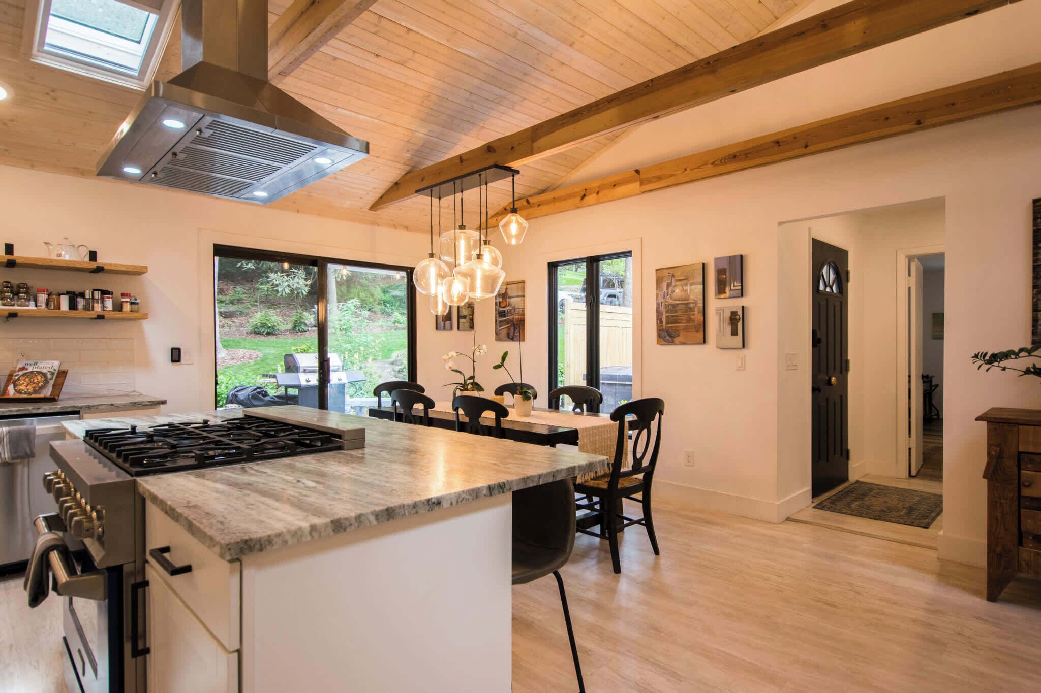 A modern kitchen with a central island, wooden beams, large windows, and a dining area with chairs and a table.