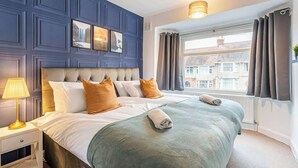 A hotel room with a large bed, a bedside table with a lamp, a window with curtains, and a view of buildings outside.
