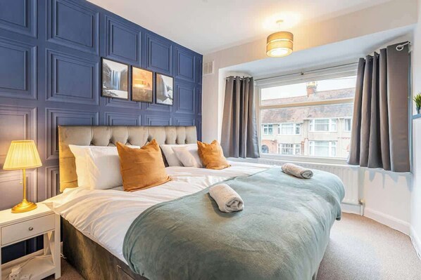 A hotel room with a large bed, a bedside table with a lamp, a window with curtains, and a view of buildings outside.
