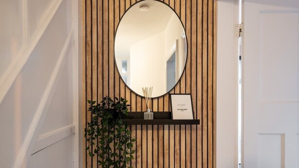 A reception area with a wooden slat wall, a round mirror, a small shelf with a plant and a framed sign, and a white door.