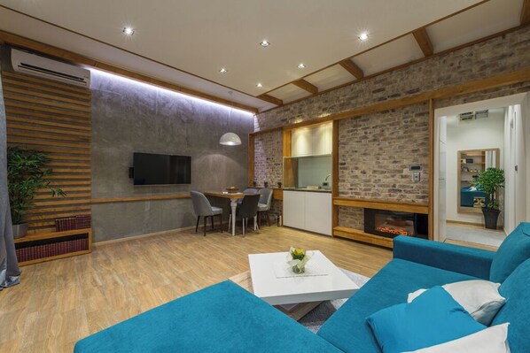 A modern living room with a fireplace, a dining area, and a kitchen with a brick wall.