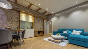 A modern living room with a blue sofa, a dining area with a table and chairs, and a kitchen with a brick wall and wooden beams. - Sapphire - Max Luxury Apartments, a Large Apartment in the Belgrade City Center (Beograd)