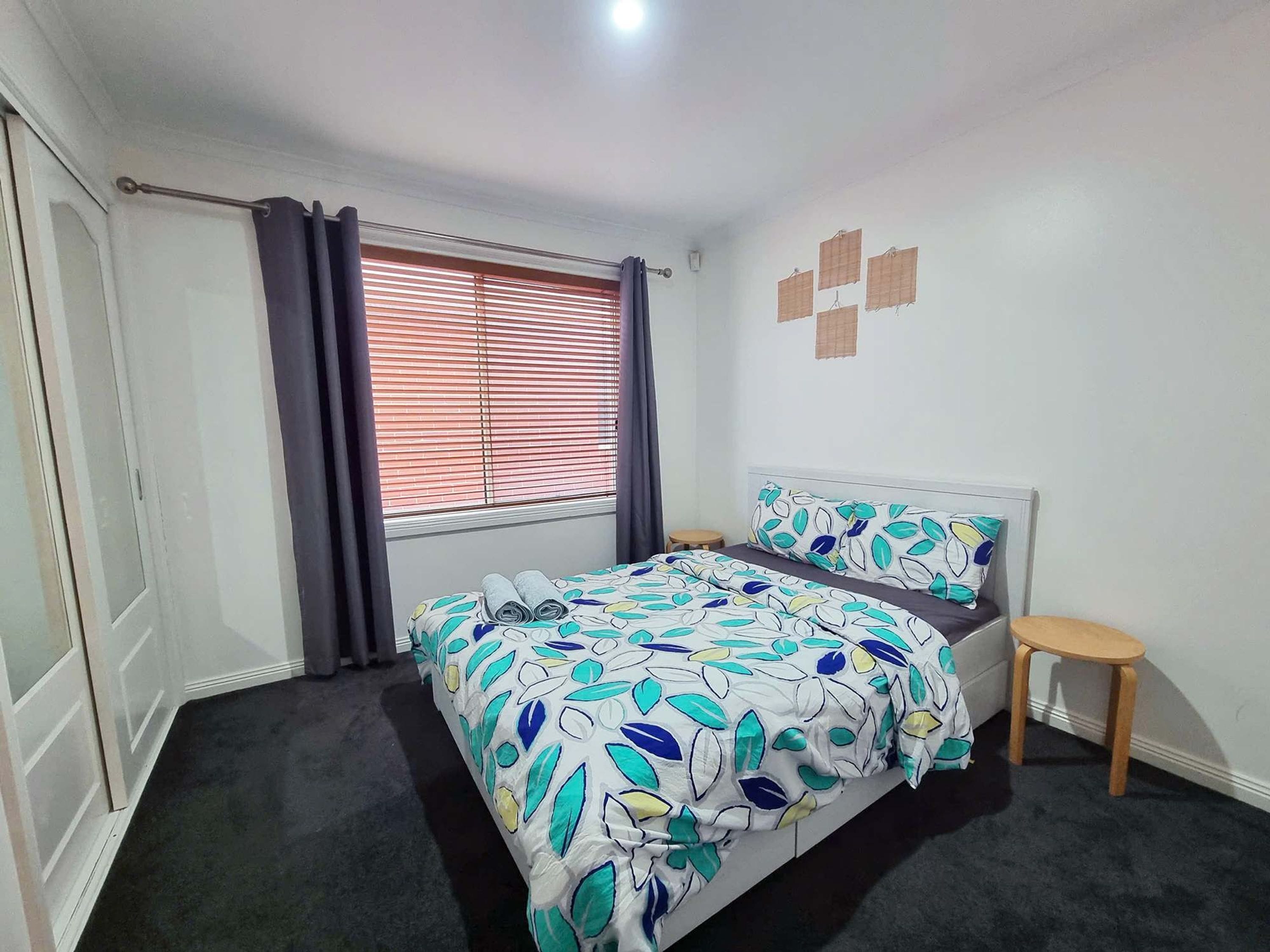 A bedroom with a bed, a small stool, a window with blinds, and a wall-mounted light fixture.