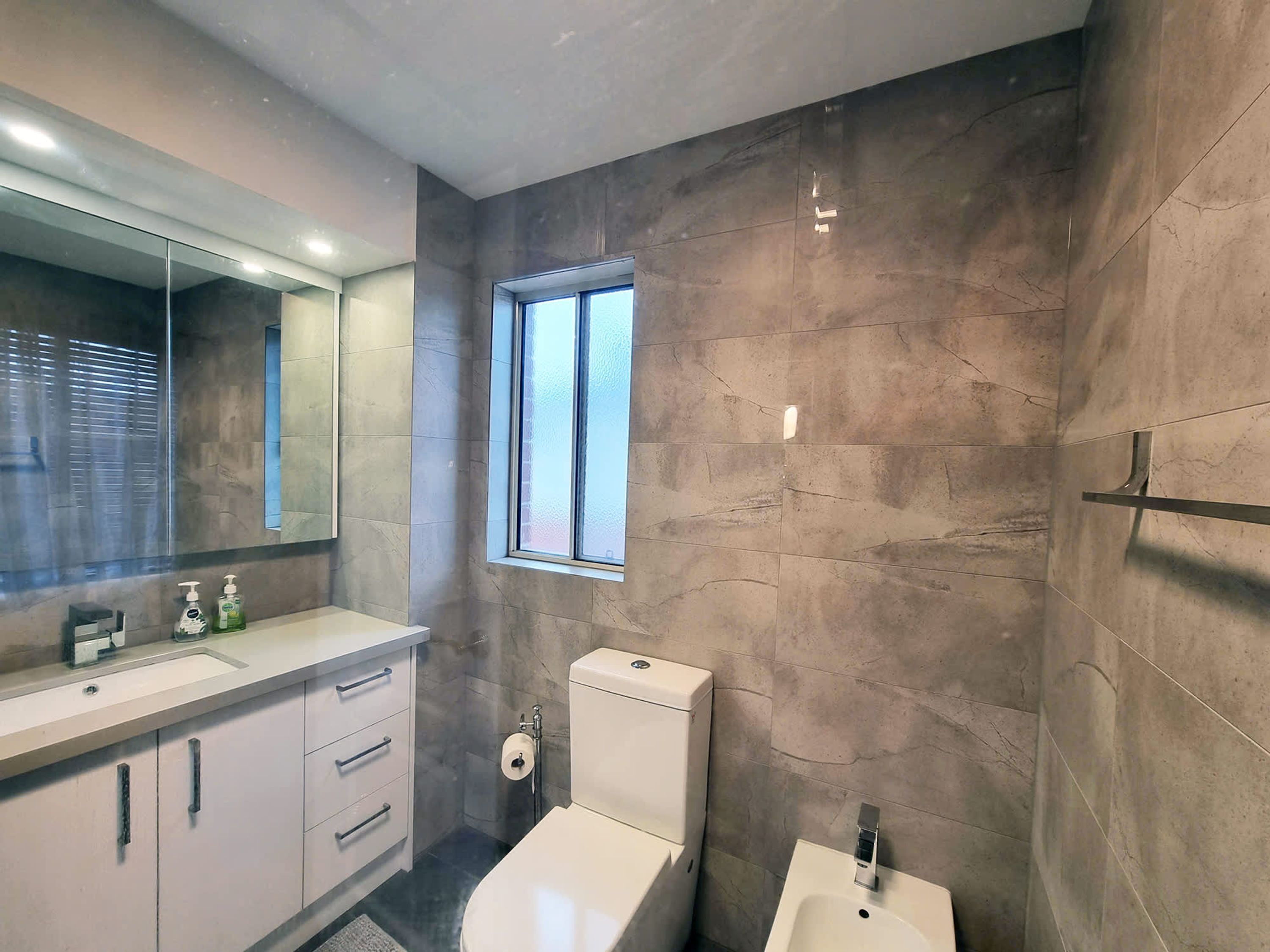A modern bathroom with a large mirror, a toilet, a bidet, and a window providing natural light.