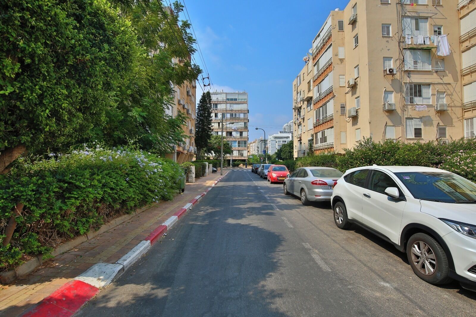 A residential street with parked cars, multi-story buildings, and greenery along the sidewalk.