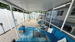 Un área de estar al aire libre con techo, que incluye un sofá azul, muebles de mimbre y una mesa de vidrio.