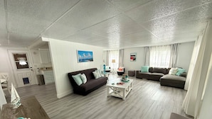 Amplia sala de estar con un sofá marrón oscuro, una mesa de centro blanca y un sofá modular gris. Hay una pequeña mesa de madera con objetos decorativos. La habitación tiene techo y paredes blancos, y una puerta que da a una zona de cocina.