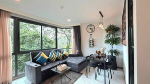 A modern living room with a grey sofa, a coffee table, and a dining area with a table and chairs. There are large windows with curtains, a wall clock, and a hanging light fixture. - A204 - 1 Bdr Condo With Sea View, Ao Nang Beach (Krabi)