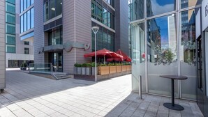 A modern building with a glass facade, outdoor seating area with red umbrellas, and a small table. - Vibrant Hub Apartment • City Centre • Balcony • Free Parking • Yoko Property (Milton Keynes)