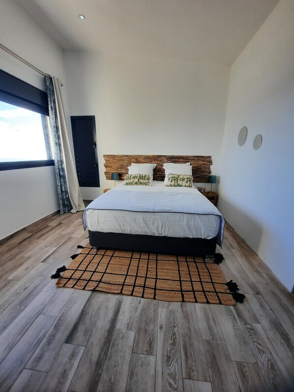 A modern bedroom with a wooden headboard, a large bed, a checkered rug, and a window with curtains. - La Villa des Oliviers (Petite-Île)