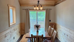 A wooden dining room with a long table and chairs, a hanging light fixture, and a window with curtains. - Riverfront Bear Cabin (Paris)