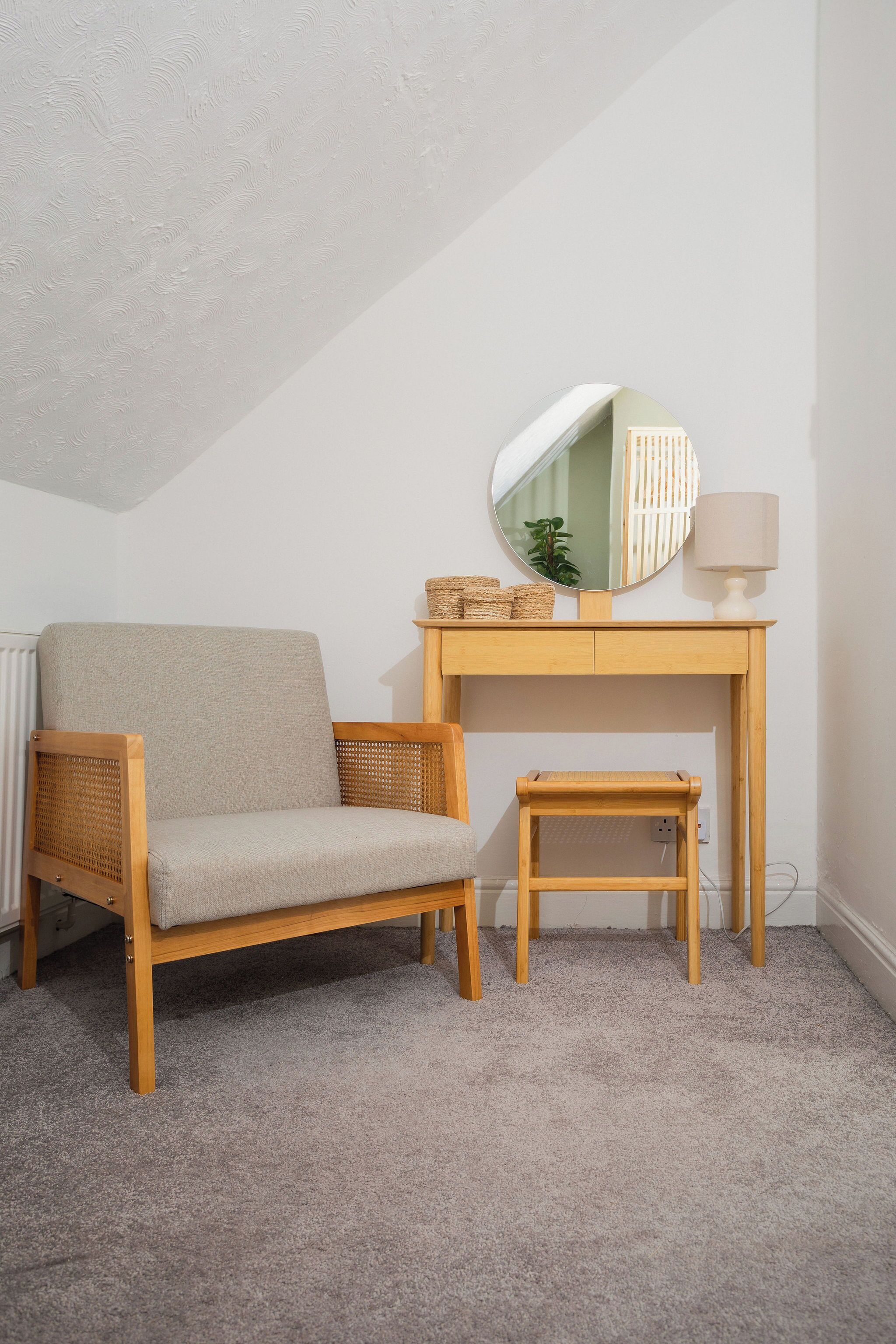 Une pièce comprenant un bureau en bois, un miroir rond, une lampe, une chaise en osier et un petit tabouret.