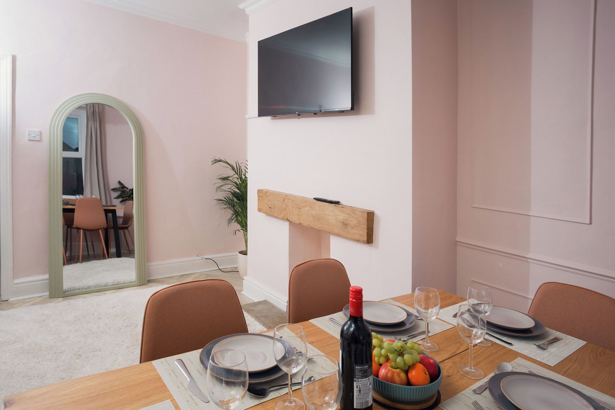 A dining room with a table set for four, a television mounted on the wall, and a mirror with an arch frame.