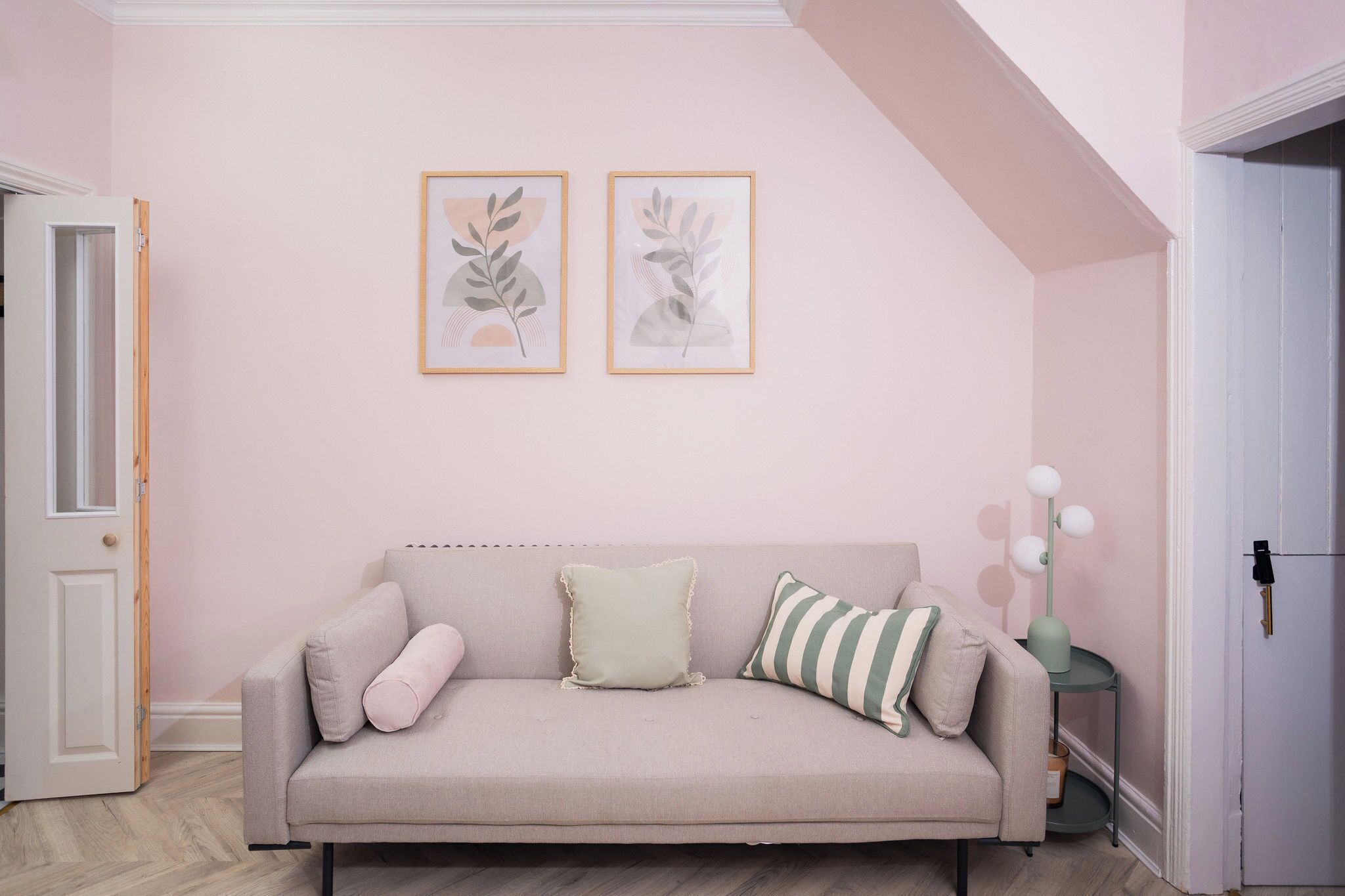 Ein Zimmer mit einem beigen Sofa, zwei gerahmten Pflanzenbildern an der Wand und einem kleinen Beistelltisch mit einer Lampe.