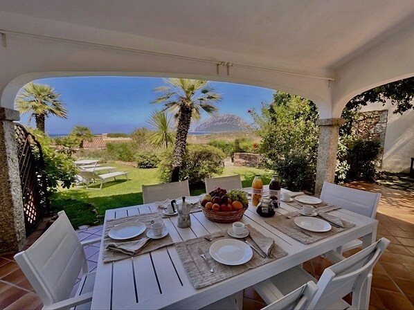 An outdoor dining area with a table set for two, overlooking a garden with palm trees and a view of the sea.