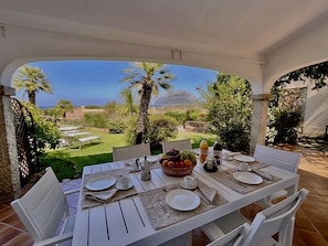 An outdoor dining area with a table set for two, overlooking a garden with palm trees and a view of the sea.