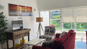 Un moderno salón con un sofá rojo, una mesa de centro de madera, un televisor y una lámpara. Hay ventanas grandes y se ve follaje verde afuera.