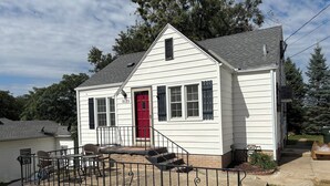 Una casa de un nivel con puerta roja, revestimiento blanco y un pequeño porche con mobiliario exterior.