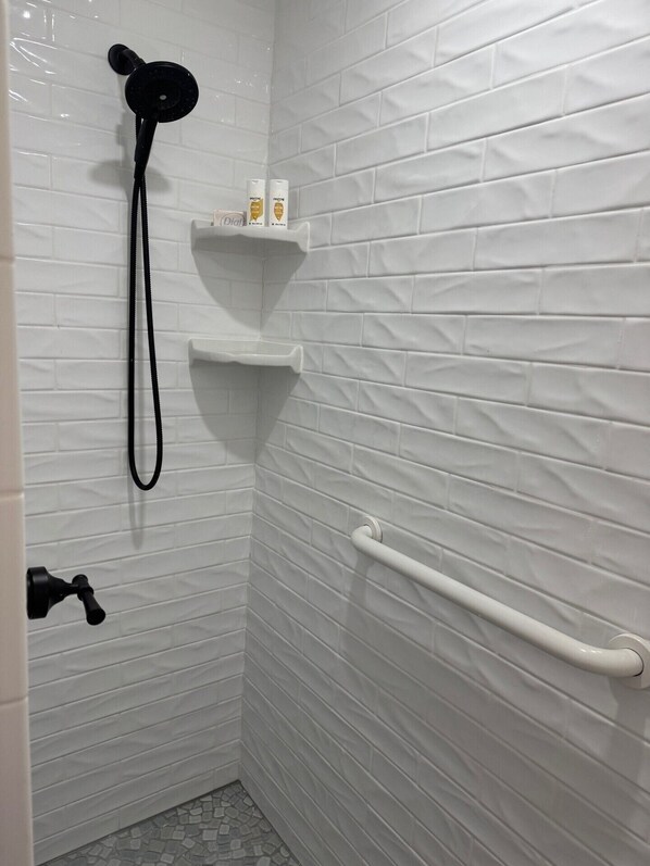 Un espace douche équipé d’un pommeau de douche à main, d’une porte-serviettes blanche et d’un distributeur de savon placé sur une étagère.