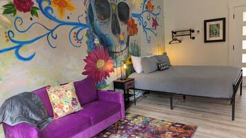 A bedroom with a bed, a purple sofa, a colorful rug, a wall mural, and a bedside table with a lamp.