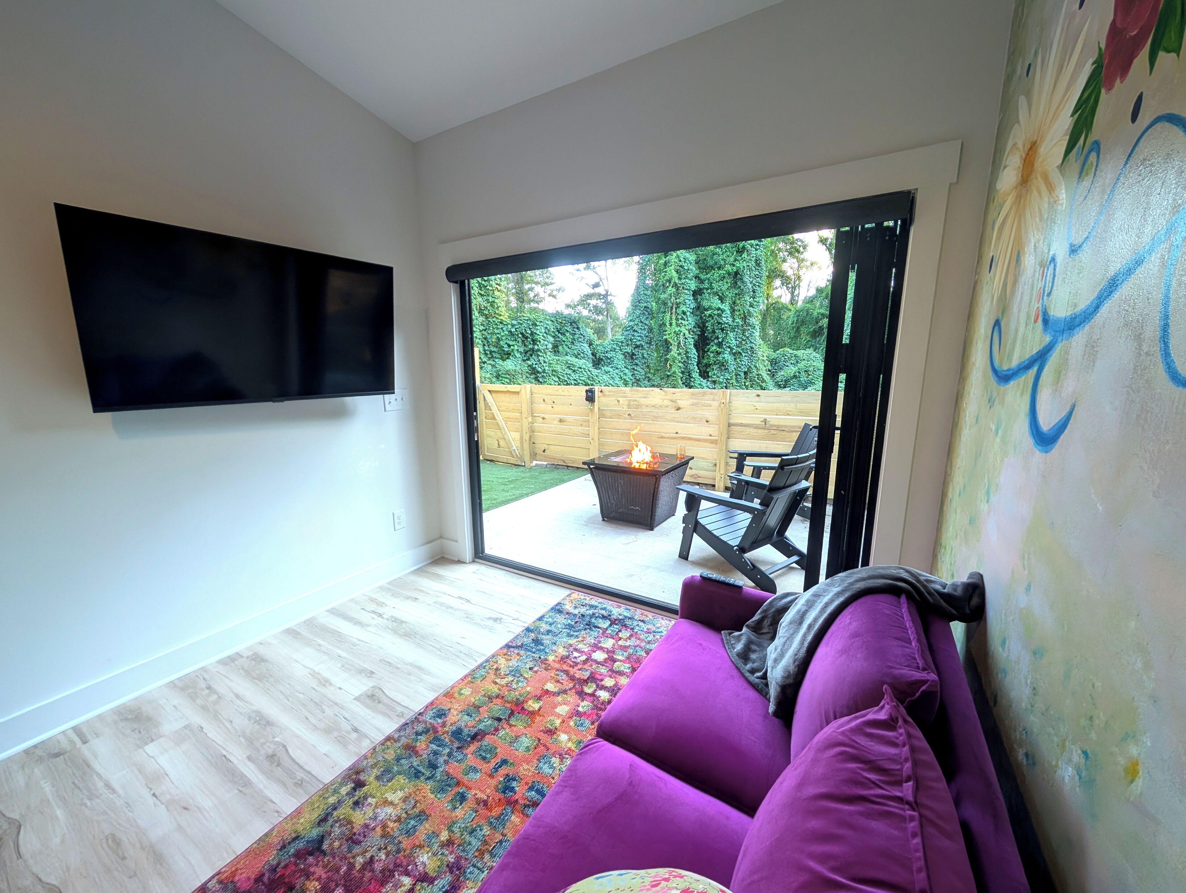 Un salon moderne avec un canapé violet, un téléviseur à écran plat fixé au mur et un tapis aux couleurs vives.