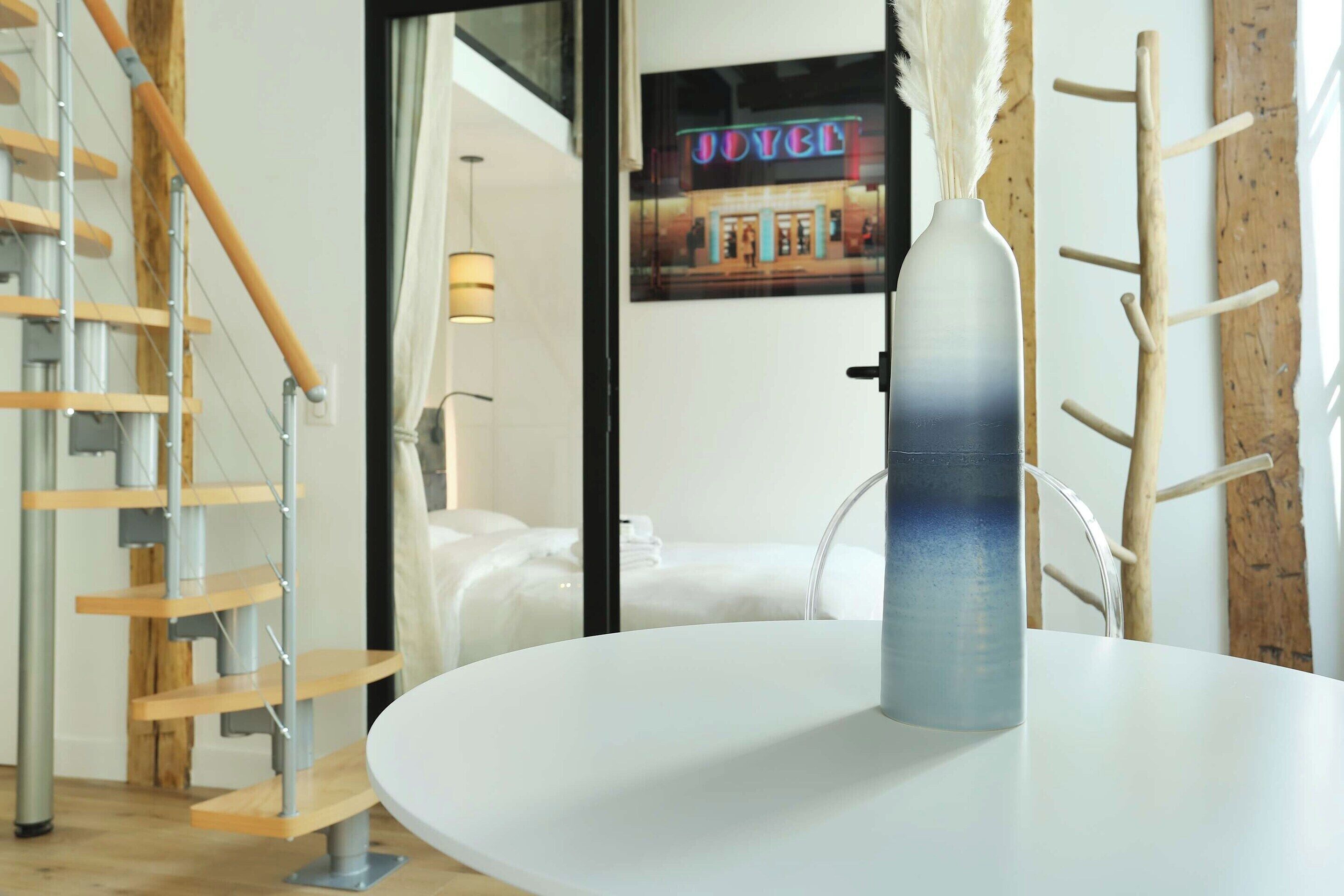 Ein modernes Hotelinterior mit einem weißen runden Tisch, einer Metallvase, einer Holztreppe und einem Bett mit weißer Bettwäsche.