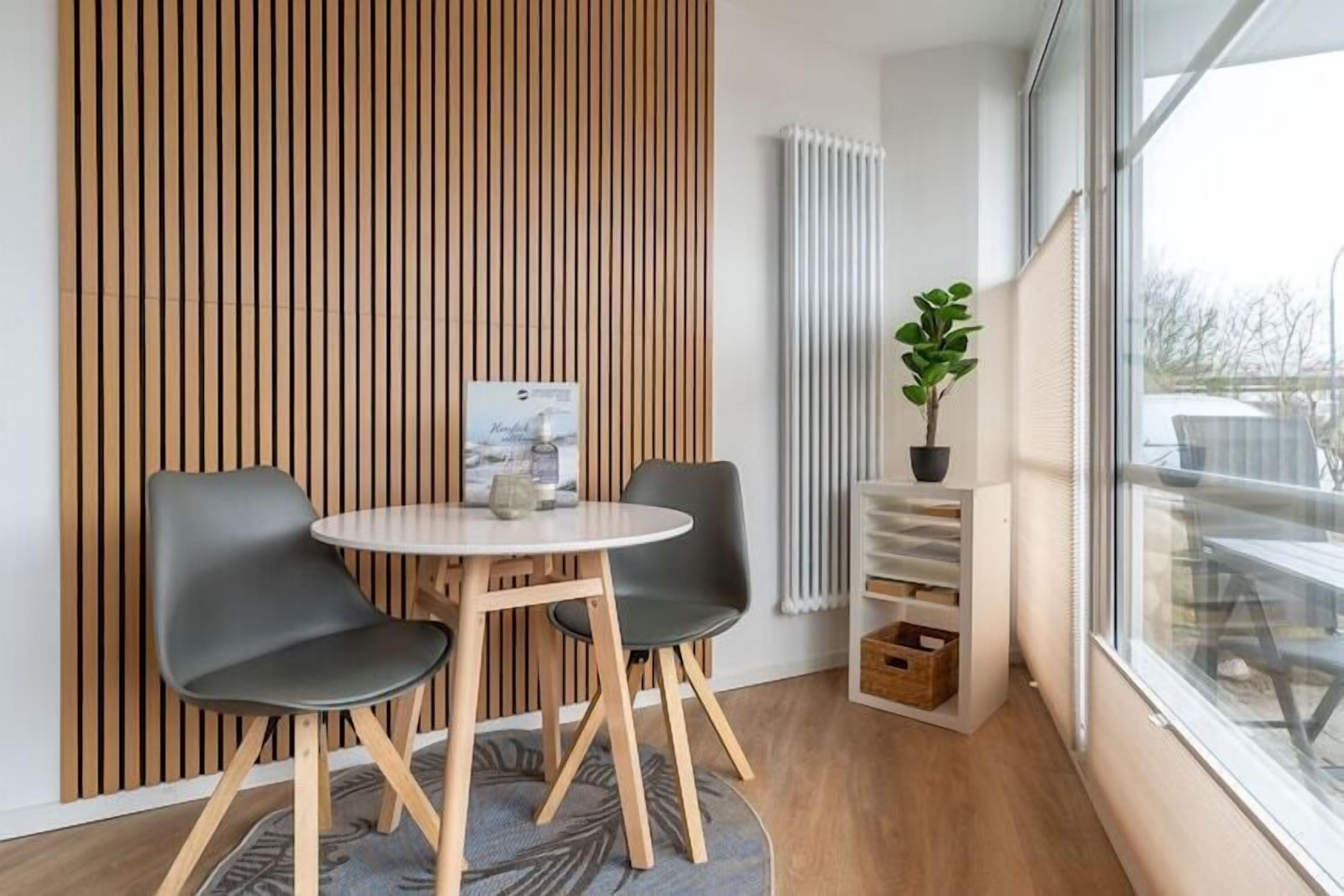 A modern room with a round table and two chairs, a wooden slat wall, a radiator, a potted plant, and a window with a view.