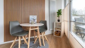 Una habitación moderna con una mesa redonda y dos sillas, un revestimiento de listones de madera, un radiador, una planta en maceta y una ventana con vistas.
