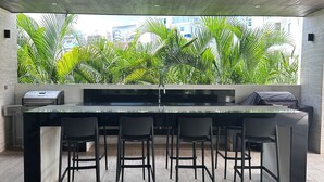 A modern bar area with a long counter, multiple stools, and a view of lush greenery and buildings through a large window. - “Elegant 3-Bedroom Apartment with Stunning Views in Cancun (Cancun)