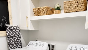 A laundry room with two front-loading washing machines, a drying rack, and shelves above holding baskets and a small plant.