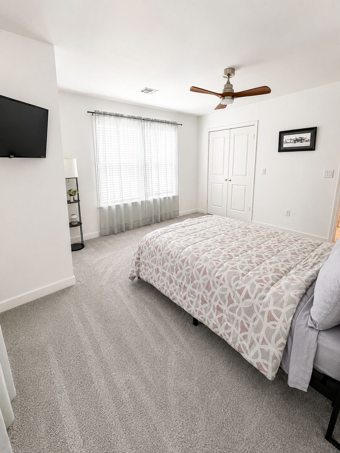 Un dormitorio con cama, ventilador de techo, ventana con cortinas, una puerta y un cuadro en la pared.