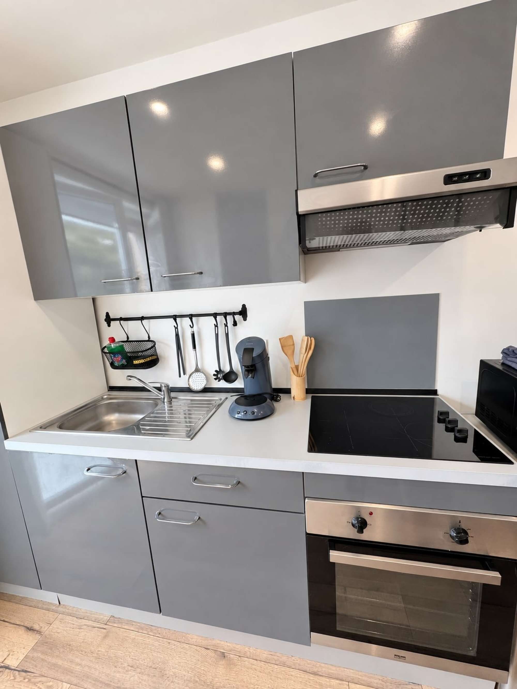 A modern kitchen with grey cabinets, a stainless steel range hood, a built-in oven, and a black stovetop.