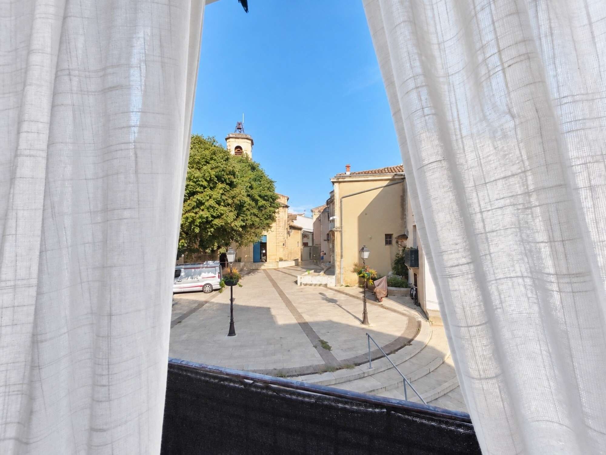 Veduta attraverso una finestra con tende bianche, che mostra una strada con edifici, un albero e un cielo azzurro limpido.