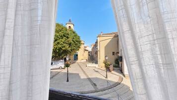 Veduta attraverso una finestra con tende bianche, che mostra una strada con edifici, un albero e un cielo azzurro limpido.