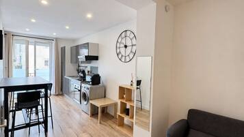 A modern living space with a dining area, a sofa, a kitchen with a washing machine, and a large wall clock.