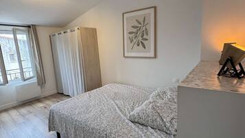 A bedroom with a bed, a window with curtains, a wall-mounted picture, and a bedside table with a lamp.