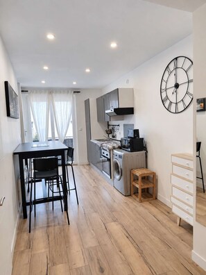 Une cuisine compacte dotée d’appareils électroménagers modernes, un coin repas avec une table et des chaises, et une grande horloge murale.