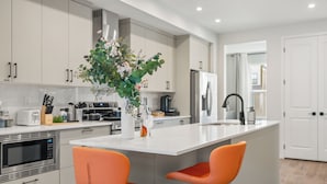 Uma cozinha moderna com armários brancos, uma ilha central com duas cadeiras laranja, uma geladeira de aço inoxidável e um micro-ondas.