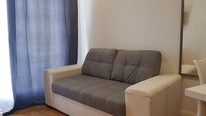 A small, single-seater sofa with a grey cushion and a white armrest, positioned against a plain wall with a window and a mirror.