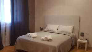 A neatly made bed with white linens and towels, a bedside table with a lamp, and a window with curtains.