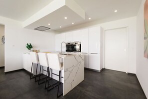 Uma cozinha moderna com ilha central, banquetas, forno embutido e armários brancos.