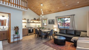 Un vaste espace de vie comprenant une table à manger, un canapé et un pouf, un plancher en bois et une vue sur l’extérieur grâce à de grandes fenêtres.