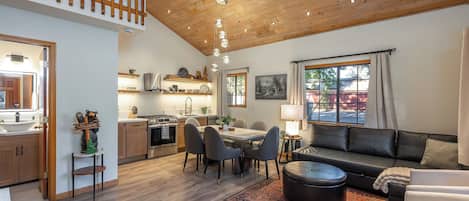 A spacious living area with a dining table, sofa, and ottoman, wooden flooring, and a view of the outdoors through large windows.