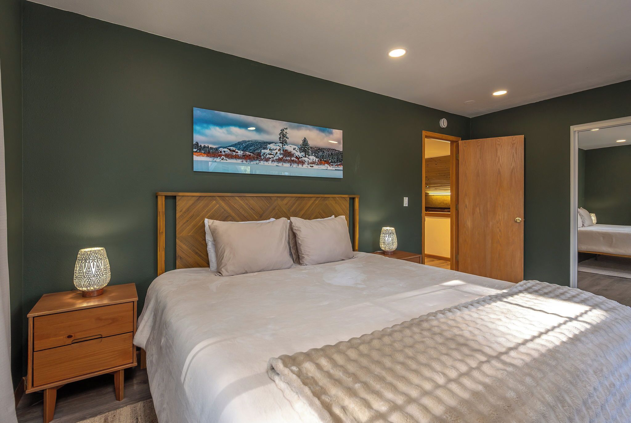 A bedroom with a large bed, a nightstand with a lamp, a wooden headboard, a painting on the wall, and a closet.
