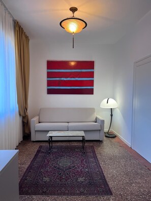 A living room with a sofa, a coffee table, a patterned rug, a wall-mounted abstract painting, and a floor lamp. - Casanova Hideaway (Venice)