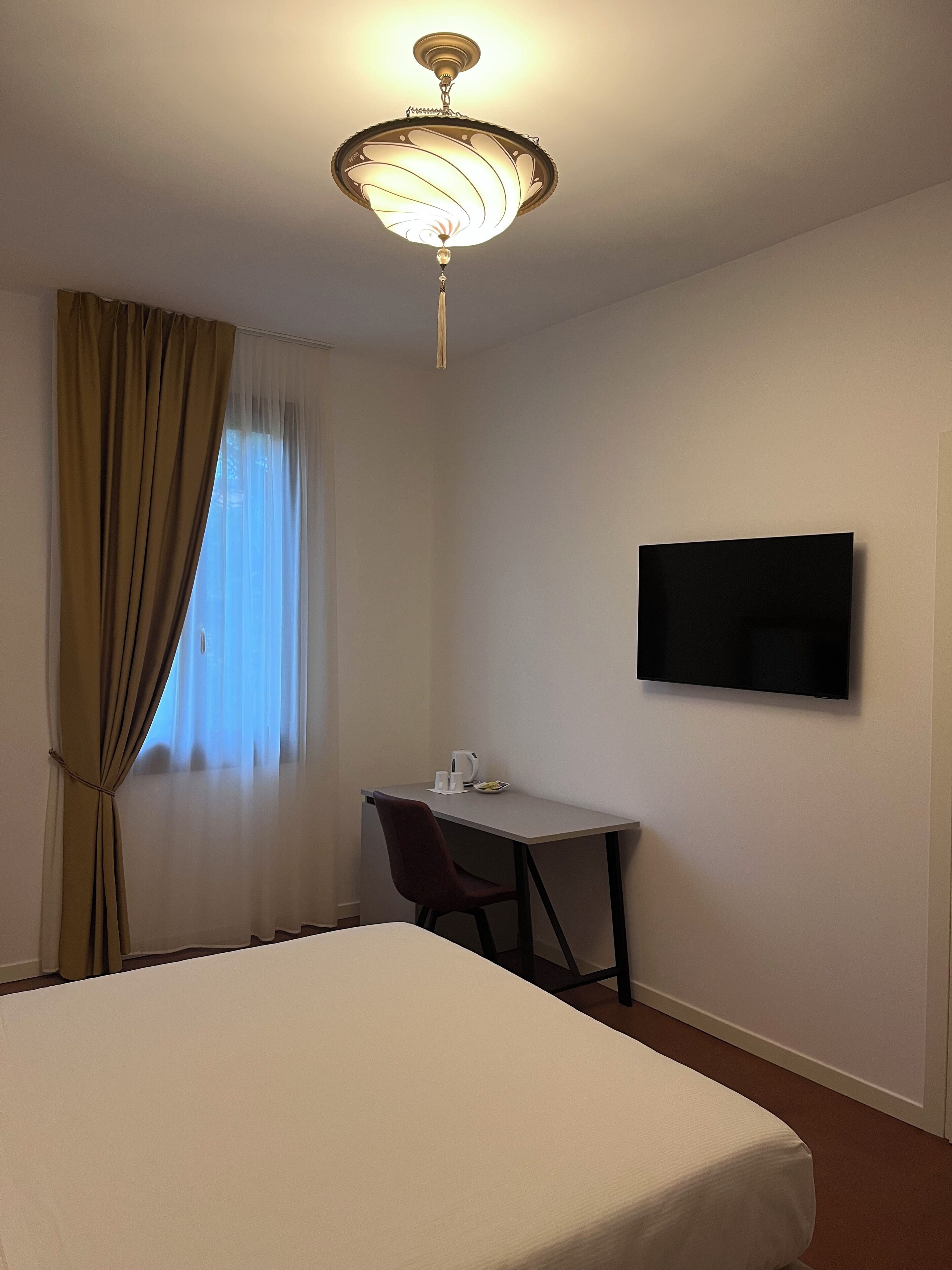 Hotellihuone, jossa on sänky, työpöytä, tuoli, televisio ja ikkuna, jossa on verhot.