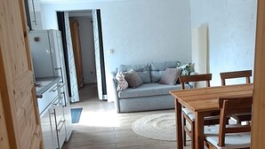 A kitchen with cabinets and a refrigerator, a dining area with a wooden table and chairs, and a gray sofa in the background. - Vacation apartment in country house style. small, cozy and quiet location (Wasserlosen)
